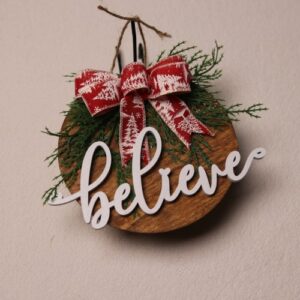 6.5 inch round sign with dark oak stain and festive wood cut cursive lettering with "believe" as a holiday message. Pine tied up with red ribbon showing white snow covered pine trees.