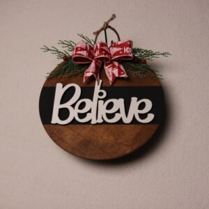 10 inch wooden circular Christmas sign with the message "Believe" in fun wood lettering cutouts. Dark walnut stained wood with a black paint stripe and white painted wood lettering. A touch of pine and holly berry tied at the top with a festive red and white bow.