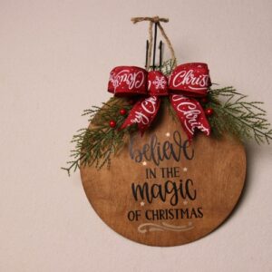 10 inch wooden circular sign with a walnut stain. Sign reads "believe in the magic of Christmas" with a whimsical font and golden stars around the words. Pine and holly berry arrangement with a festive red and white Christmas bow.