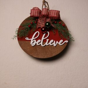 6.5 inch round wooden Christmas sign in walnut stain with a red paint stripe and cursive white wood cutout lettering. "Believe" in the Christmas magic. Complete with festive metallic jingle bells, pine, and holly berries tied together with a checkered red and white bow.