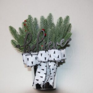 Small table-top holiday decoration. Metal pale full of faux pine needles and holly berries. Fun wood cutout word "believe" in festive font. Complete with holiday festive white bow featuring pine trees and cross stitching edges.