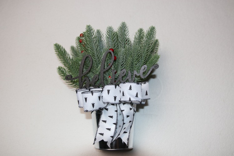 Small table-top holiday decoration. Metal pale full of faux pine needles and holly berries. Fun wood cutout word "believe" in festive font. Complete with holiday festive white bow featuring pine trees and cross stitching edges.