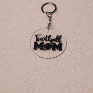 Key chain for the ultimate Football Mom. Clear circular disc keychain with fun "Football Mom" text. Features a football graphic as the "O" in "MOM."