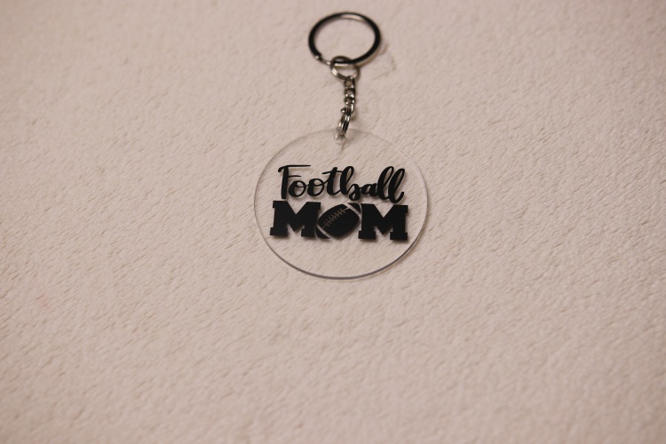 Key chain for the ultimate Football Mom. Clear circular disc keychain with fun "Football Mom" text. Features a football graphic as the "O" in "MOM."