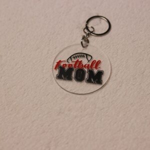 Key chain for the ultimate Football Mom. Clear circular disc keychain with fun "Football Mom" text. Features a football graphic and Colby-Jack's home town colors of red and black.