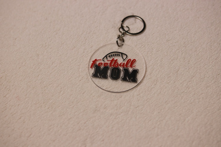 Key chain for the ultimate Football Mom. Clear circular disc keychain with fun "Football Mom" text. Features a football graphic and Colby-Jack's home town colors of red and black.
