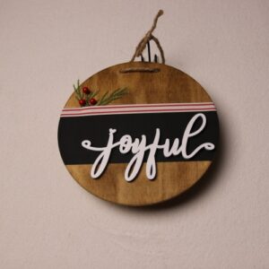 6.5 inch round wooden sign with dark oak stain. Cute cursive wood cut lettering wishing a "joyful" holiday season. A splash of colors with pine and holly berries peaking out of a small white and red pin-striped ribbon.