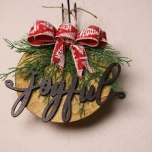 6.5 inch round wooden sign with oak stain. Cute cursive wood cut lettering wishing a "joyful" holiday season. A splash of color with pine tied together with a red ribbon forested with white pine trees.