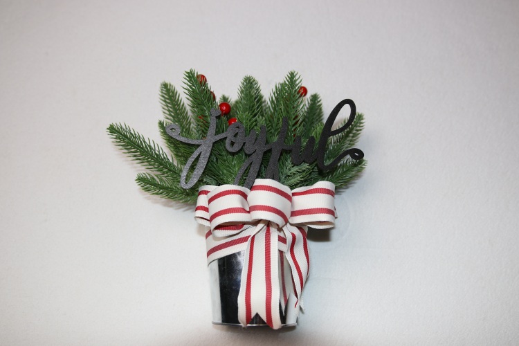 Small table-top holiday decoration. Metal pale full of faux pine needles and holly berries. Complete with wooden lettering cutout with the message "joyful" above a festive red and white Christmas ribbon.