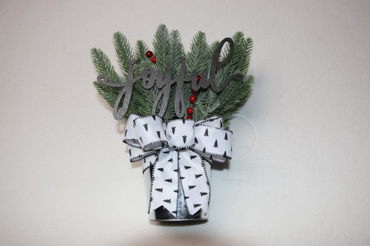 Small table-top holiday decoration. Metal pale full of faux pine needles and holly berries. Complete with wooden lettering cutout with the message "joyful" above a festive white and black Christmas ribbon featuring little pine trees.