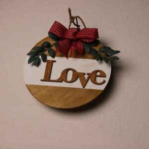 10 inch wooden circular sign in light oak stain. White paint stripe under wooden letter cutouts in medium oak stain spelling "Love." Small green leaves tied together with pin-striped red and white bow for a splash of color.