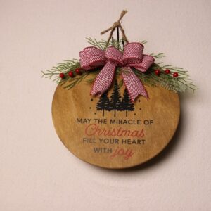 10 inch wooden circular sign with an oak stain with green and red Christmas lettering. Festive pine tree graphics and a warm Christmas greeting under a touch of pine and holly berry tied together by a checkered white and red bow.