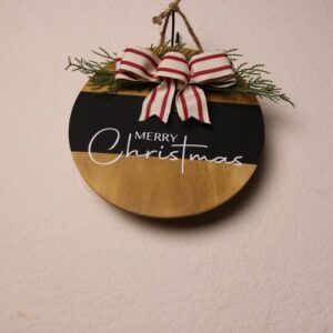 10 inch wooden circular sign with an oak stain, a black painted stripe and white lettering wishing visitors a "Merry Christmas." Festive warm Christmas greeting under a touch of pine tied together by a checkered white and red bow.