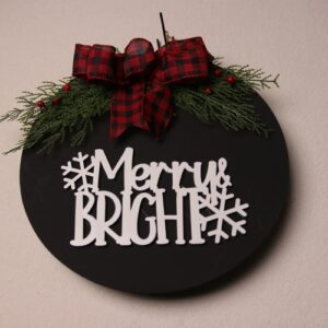 14 inch circular wooden sign, painted black with white painted wood cutout lettering featuring "Merry & Bright" with some snow flakes, pine and holly berry. Tied off with a red and black bow.