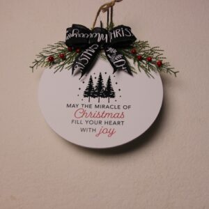 10 inch round sign painted white with green pine trees and festive green and red lettering featuring a warm Christmas message. Complete with pine and holly berry foliage tied together with a black and white Christmas bow.