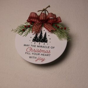 10 inch wooden circular sign painted white with green and red Christmas lettering. Festive pine tree graphics and a warm Christmas greeting under a touch of pine and holly berry tied together by a checkered white and red bow.