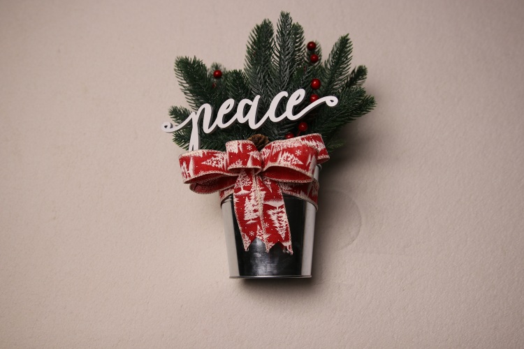 Small table-top holiday decoration. Metal pale full of faux pine needles and holly berries. Complete with cute red and white ribbon adorned with Christmas tree and snow flake pattern. Festive wood lettering reading "peace" popping out of the pines.
