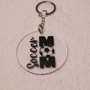 Key chain for the ultimate Soccer Mom. Clear circular disc keychain with fun "Soccer Mom" text. Features a soccer ball graphic as the "O" in "MOM."