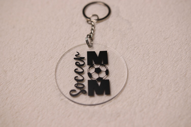 Key chain for the ultimate Soccer Mom. Clear circular disc keychain with fun "Soccer Mom" text. Features a soccer ball graphic as the "O" in "MOM."