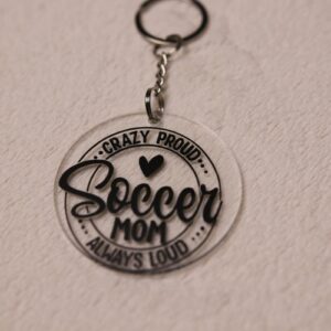 Key chain for the ultimate Soccer Mom. Clear circular disc keychain with fun "Soccer Mom" text. Features a heart graphic and the text "Crazy Proud Soccer Mom Always Loud"