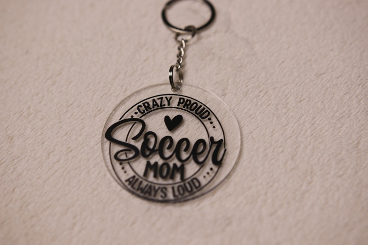 Key chain for the ultimate Soccer Mom. Clear circular disc keychain with fun "Soccer Mom" text. Features a heart graphic and the text "Crazy Proud Soccer Mom Always Loud"