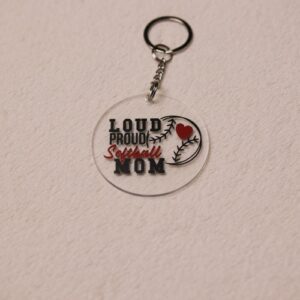 Key chain for the ultimate Softball Mom. Clear circular disc keychain with fun "Softball Mom" text. Features a heart and baseball graphic and the text "Loud Proud Softball Mom"