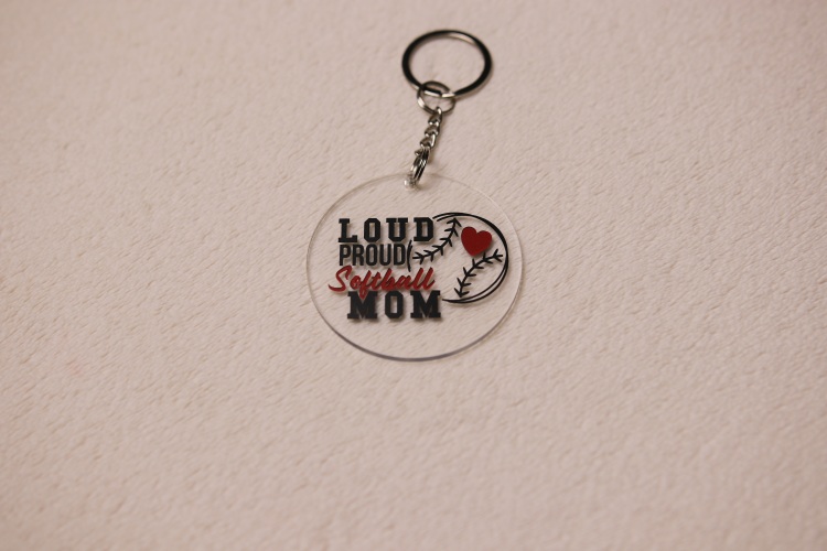 Key chain for the ultimate Softball Mom. Clear circular disc keychain with fun "Softball Mom" text. Features a heart and baseball graphic and the text "Loud Proud Softball Mom"