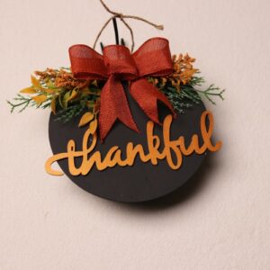 6.5 inch round wooden sign painted black to make the orange wood-cut lettering pop. Cute and playful cursive wood lettering reminding family, friends, and guests to be "thankful" this Thanksgiving season. Additional splashes of color from pine and fall foliage tied with a crimson ribbon.