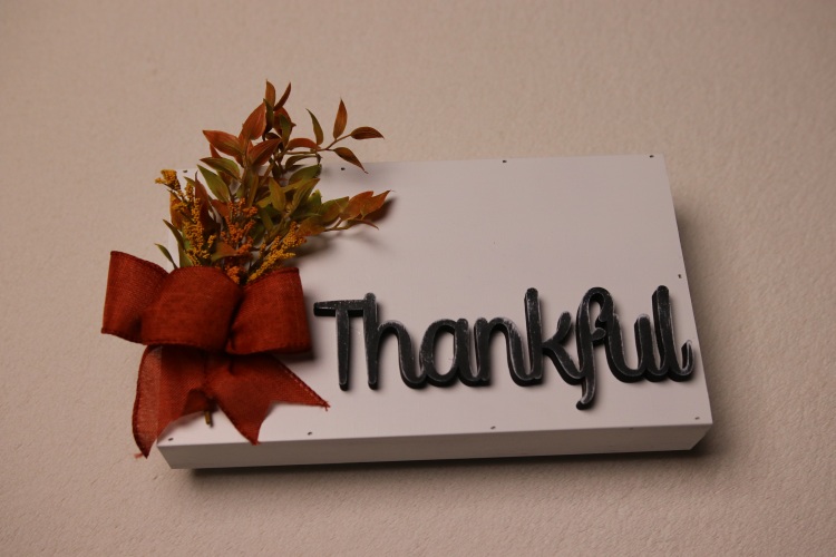 Box sign for fall decorating. Thanksgiving decoration featuring a white painted box sign with the wood lettering cutout "Thankful." Rustic sign look with distressed lettering and pops of fall colors with faux plants tied together with a crimson red/orange fall colored bow.