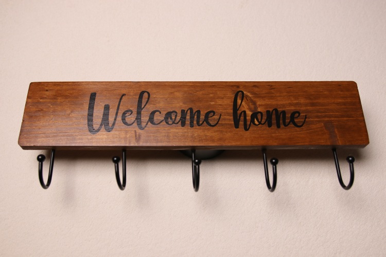 "Welcome home" coat hanging sign in dark oak stain. Five coat hanging hooks in black metal.