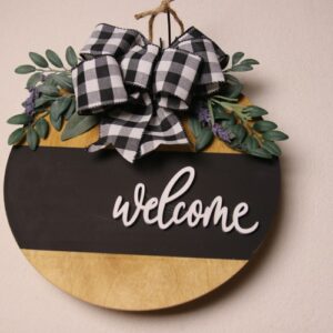14 inch wooden circular sign with light oak stain, black painted stripe and wooden cutout cursive lettering. Featuring the greeting "Welcome." Adorned with black and white lettering with leaves and lavender foliage.