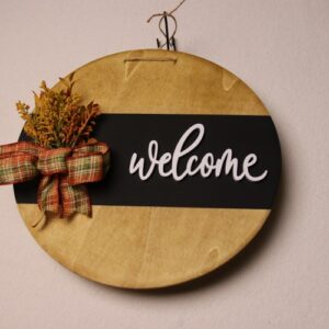 Large circular wooden sign with light oak stain and black painted stripe. White wooden cutout cursive lettering. Fall colored ribbon tied around fall foliage.