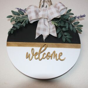 14 inch wooden circular welcome sign. White and black paint with a touch of light oak stain and cursive wood lettering cutouts. A touch of small leaves and lavender tied together with a white checkered bow.