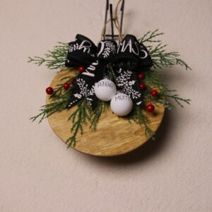 6.5 inch round wooden sign with light oak stain. "Believe" and "Merry" displayed on cute mini white Christmas ornaments hanging from a black bow with white "Christmas" lettering. Completed with pine and holly berries and a rustic twine cord for hanging.