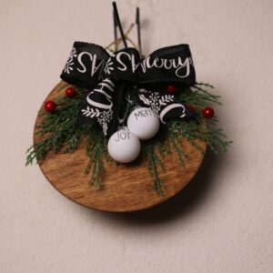 6.5 inch round wooden sign with walnut stain. Cute mini Christmas ornaments with "MERRY" and "JOY" in festive fonts. A splash of colors with pine and holly berries tied together with a black bow showing a white Christmas message.