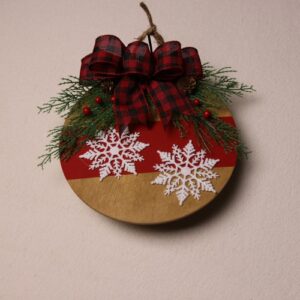 6.5 inch round wooden sign with oak stain. Cute and playful large white snowflakes across a painted red stripe. Complete with a splash of pine and holly berry tied with a big checkered red and black bow.